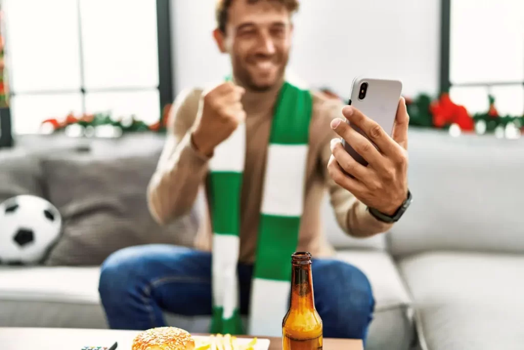 Homem branco comemorando vitória em aposta online com celular na mão